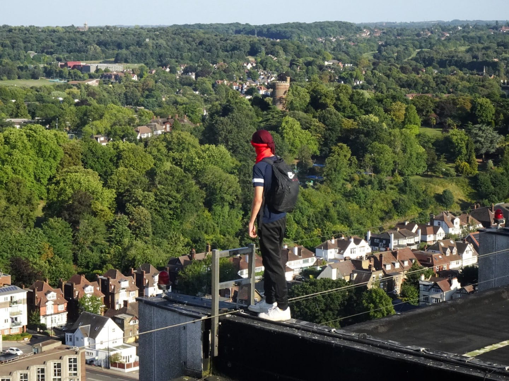Person in red cap and dark clothing walking on tightrope high above suburban neighbourhood with houses and green trees below.