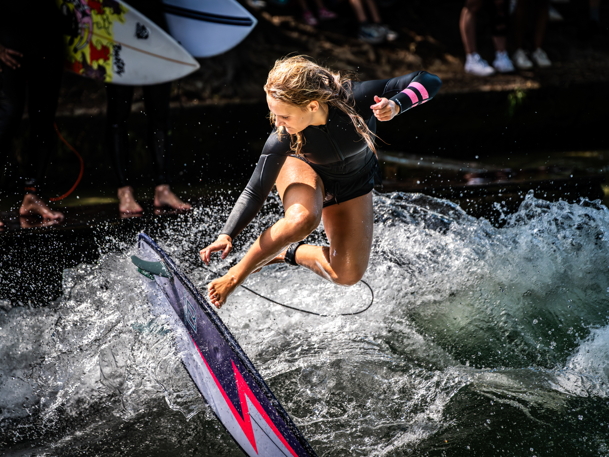 Woman wakesurfing behind boat, crouched on black and pink surfboard with water splashing around her.