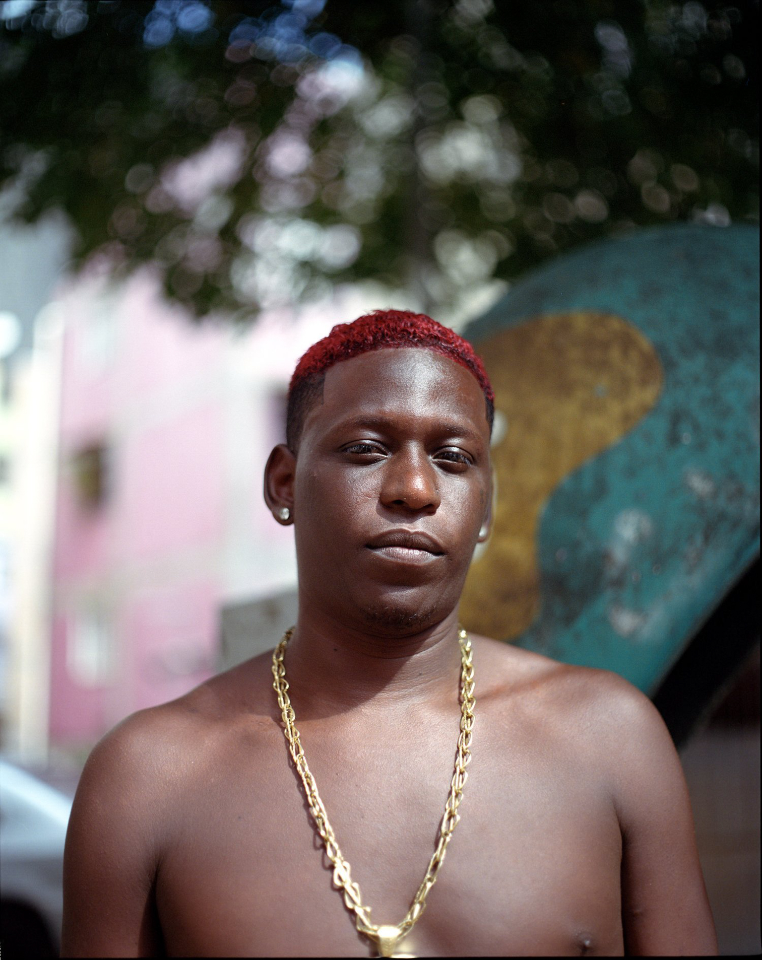 Shirtless Black man with red-dyed hair and gold chain necklace, blurred colourful buildings and foliage in background.