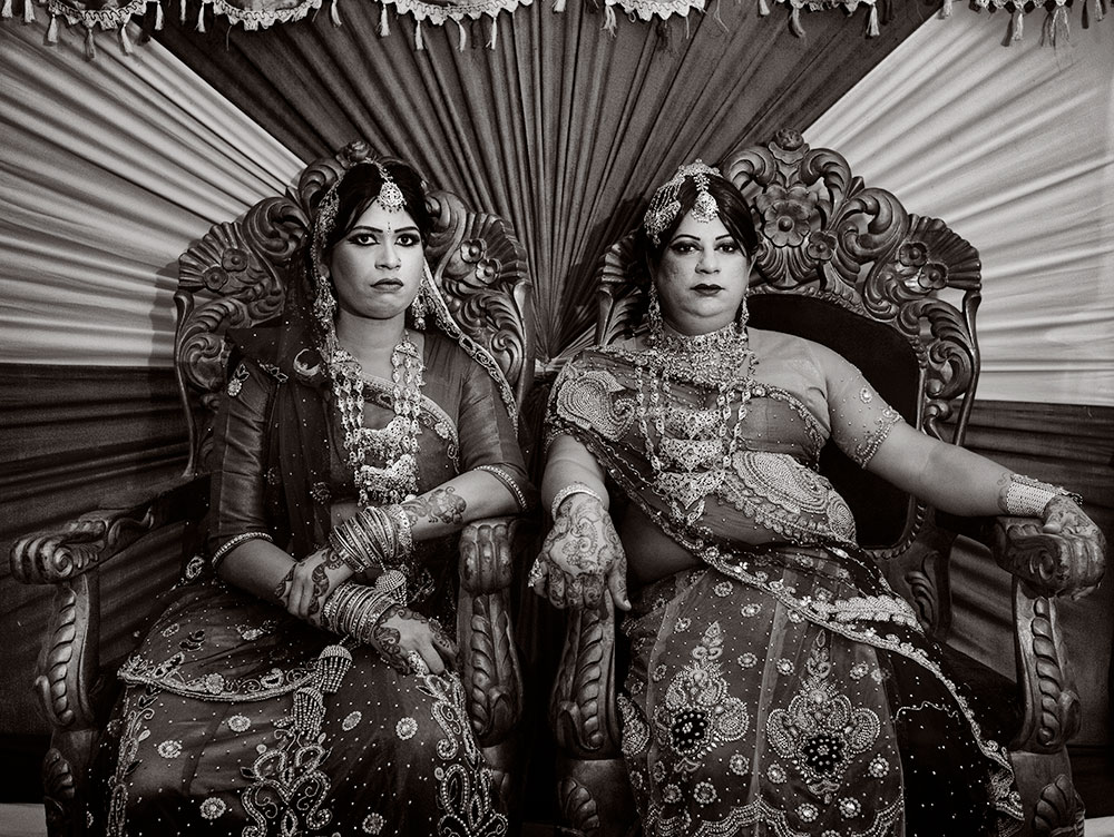 Two women in ornate traditional dress sitting on carved wooden chairs against pleated fabric backdrop with decorative fringe border.