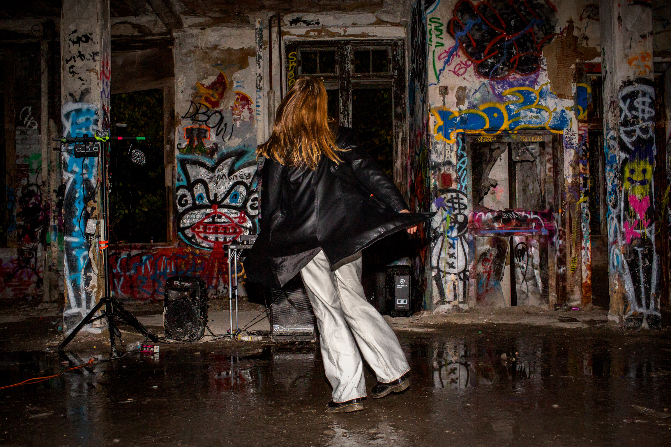 Person in black jacket and white trousers walking through graffiti-covered abandoned building with colourful street art on walls.