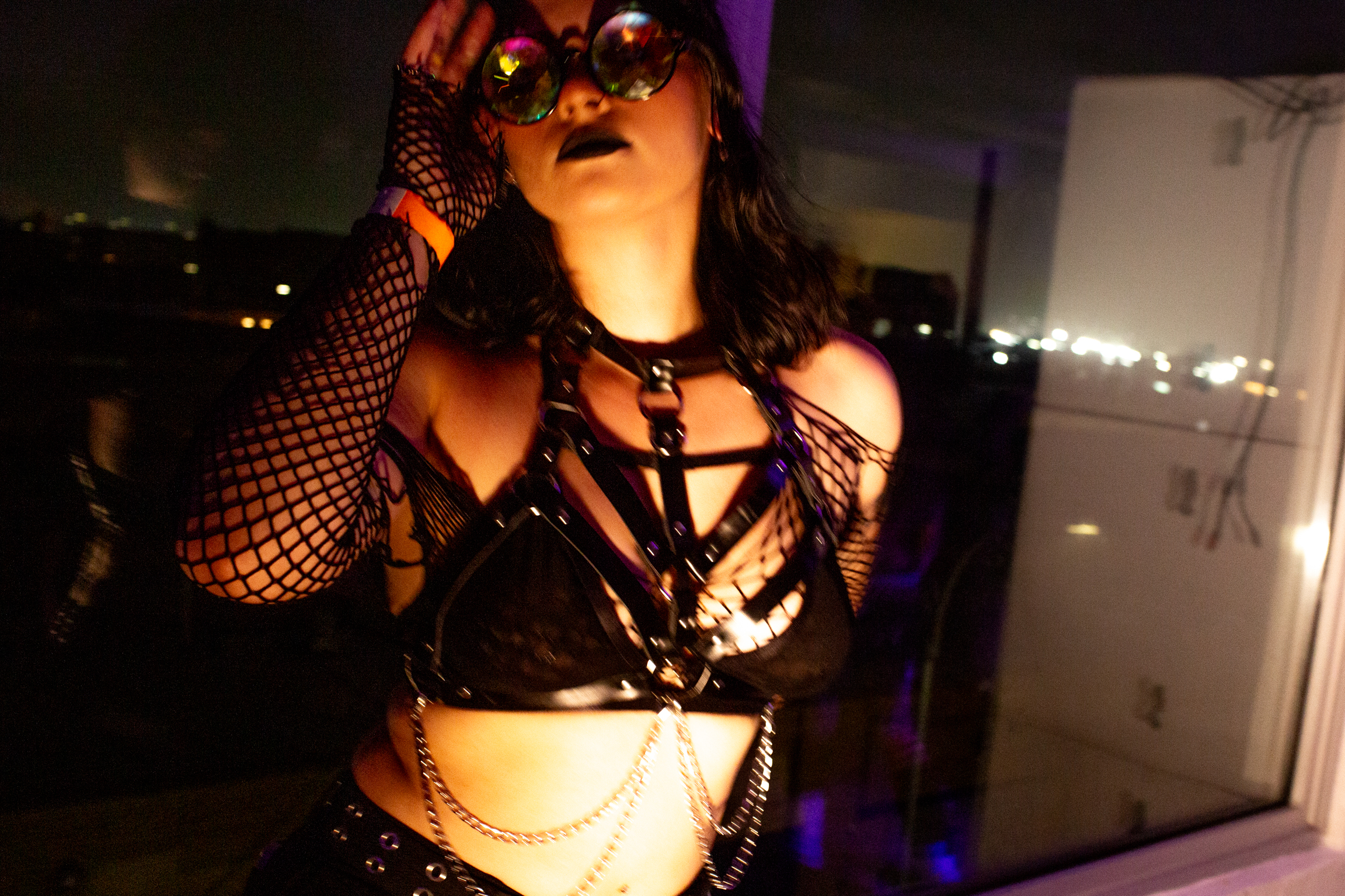 Woman in black leather harness top and fishnet gloves with reflective sunglasses, purple and orange lighting against urban night backdrop.