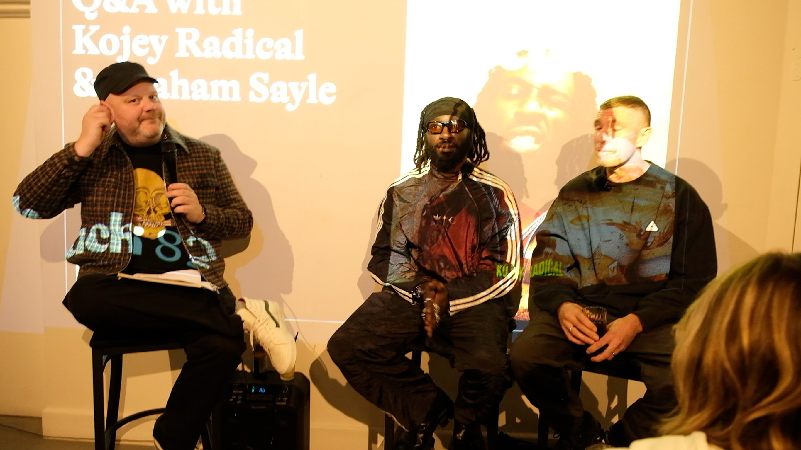Three men seated on stage during panel discussion, projection screen behind showing event title, audience member visible in foreground.