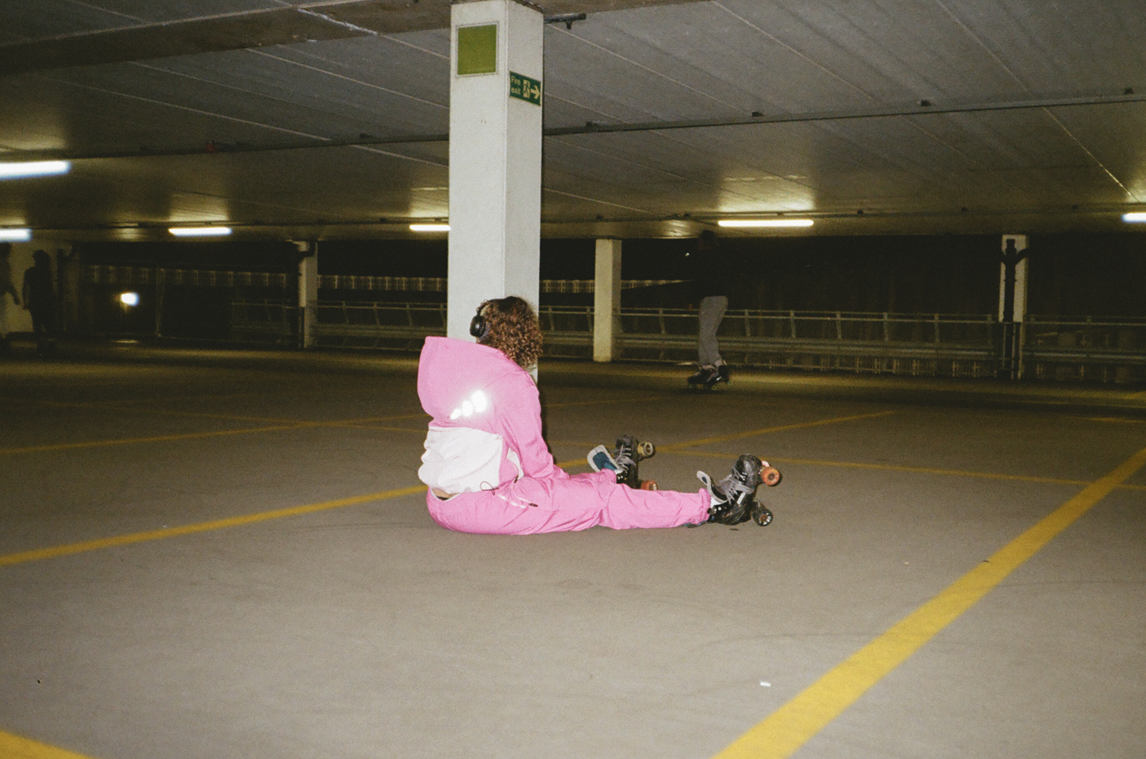 Person in bright pink jacket sitting on concrete floor of underground car park with white pillars and yellow lines.