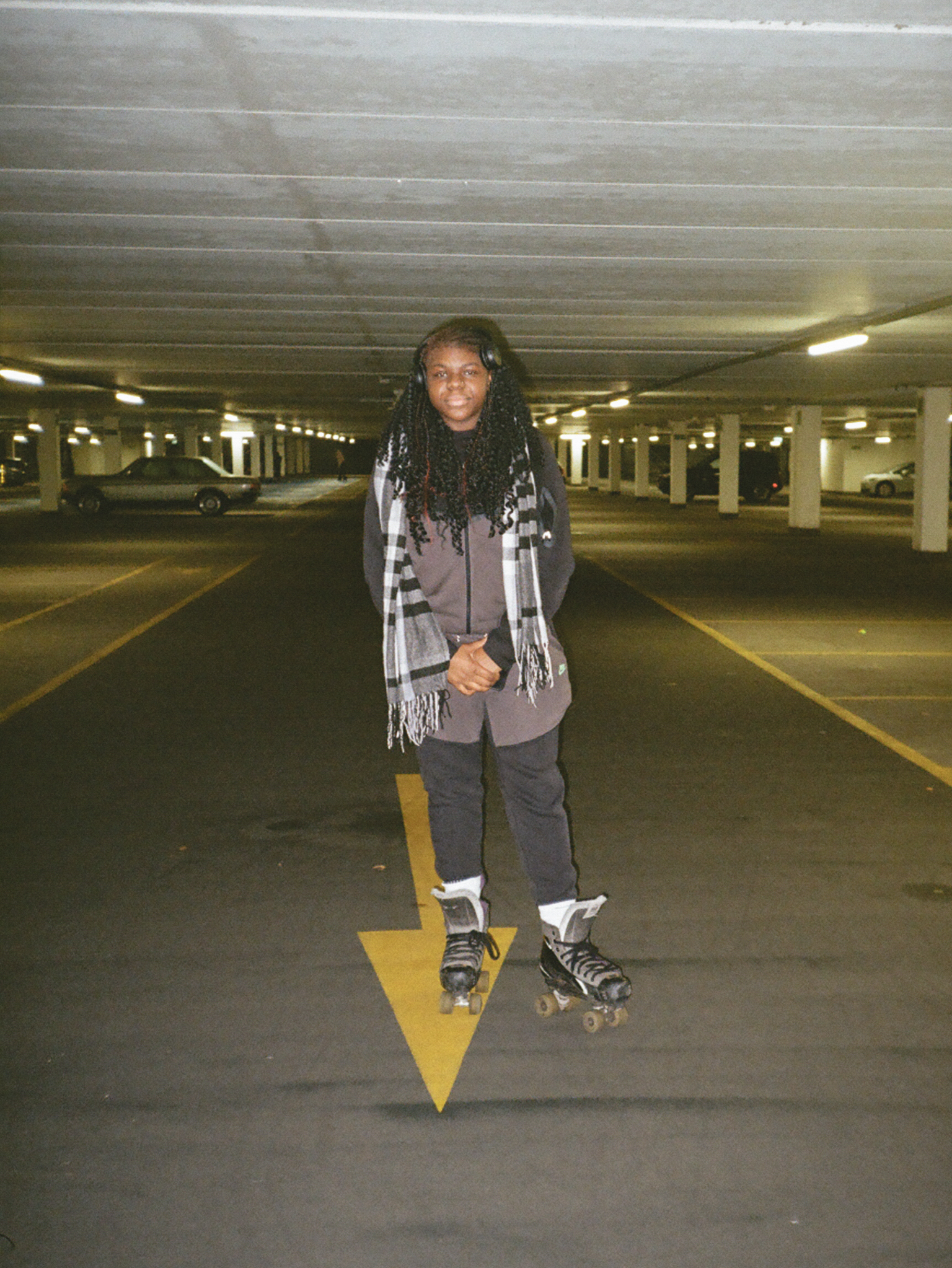 Person with dreadlocks wearing grey hoodie and patterned jacket stands on rollerblades in concrete car park with yellow arrow marking.