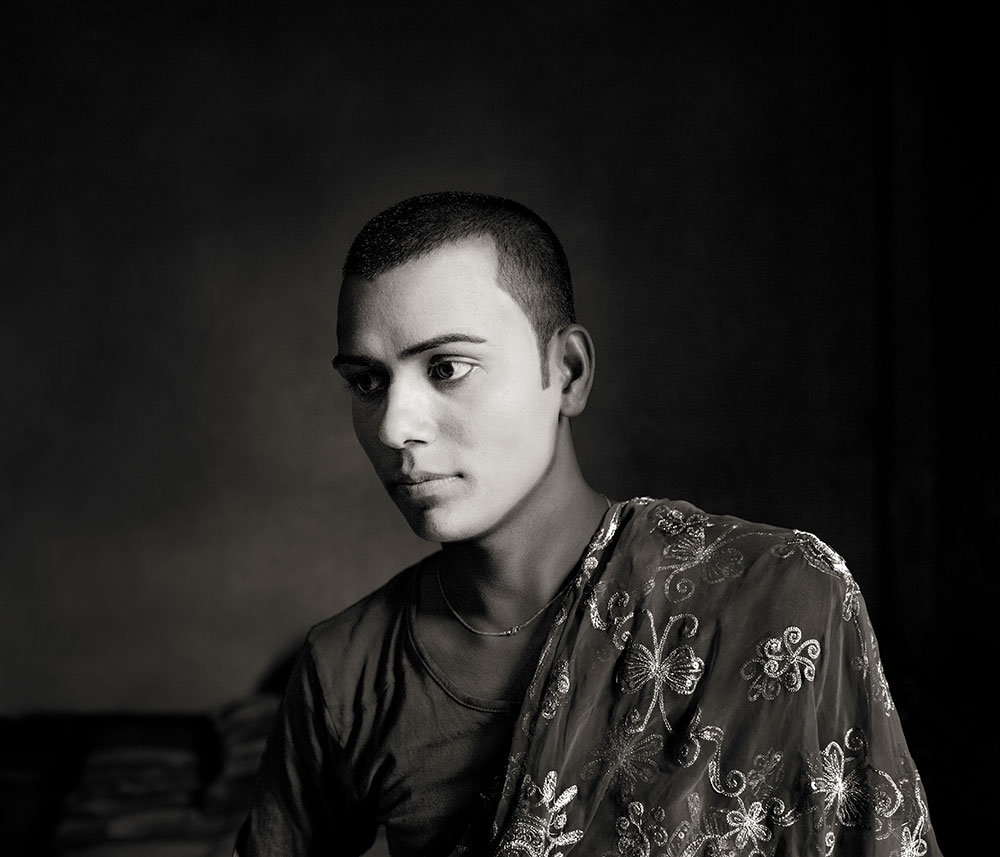 Young man with short dark hair wearing olive green patterned shirt, looking to the left against dark grey background.