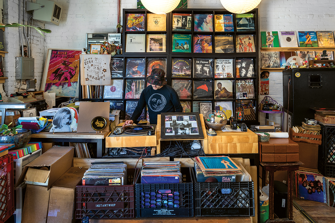 Record shop interior with vinyl albums displayed on wall shelves and in crates, person browsing at counter, warm lighting.