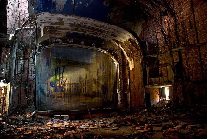 Dilapidated interior of an old theatre with ornate archways, damaged floors, and a large projection screen in the centre.