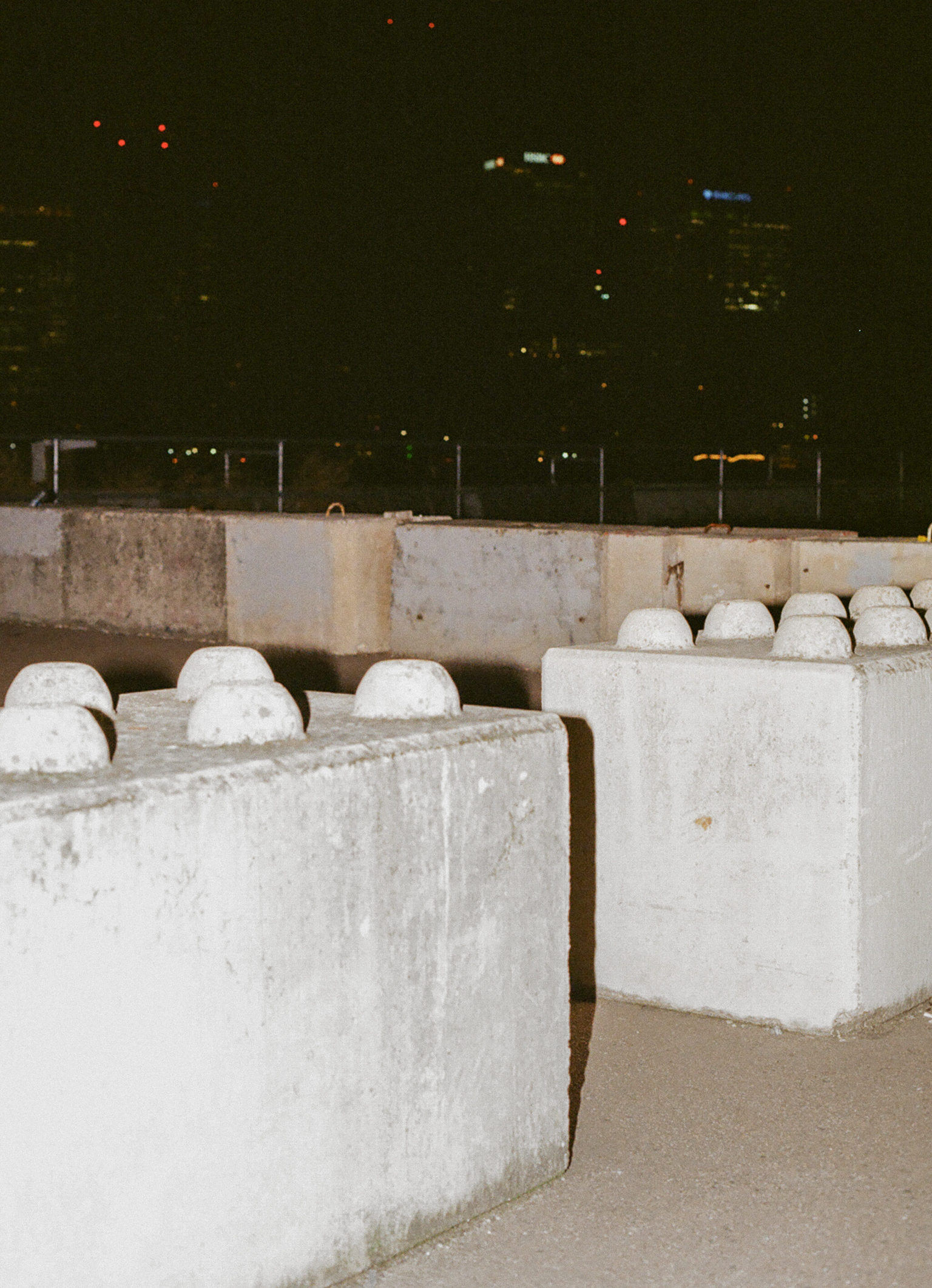 Large concrete blocks with rounded tops arranged in rows, city lights visible in dark background beyond concrete barrier.