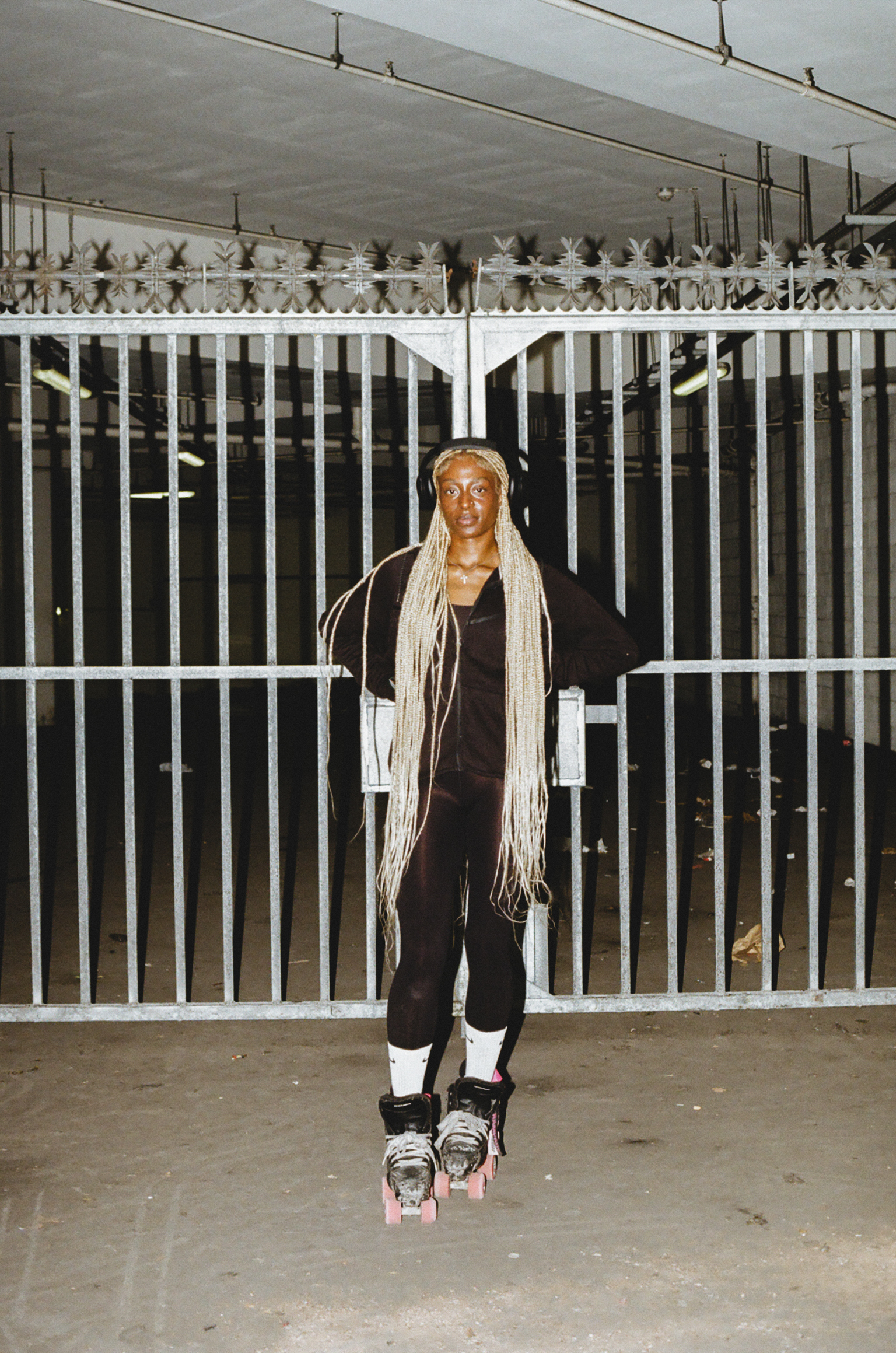 Person with long blonde hair wearing black outfit and rollerskates, standing in front of white metal gate with decorative spikes.