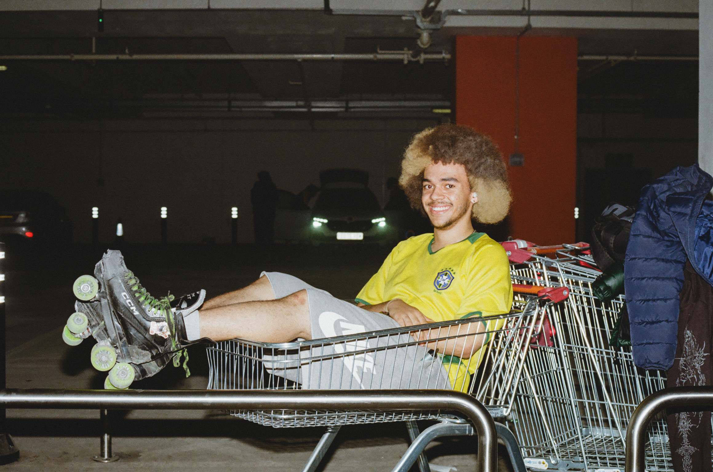 Person in yellow shirt and roller skates sitting in shopping trolley in car park, legs extended, smiling at camera.