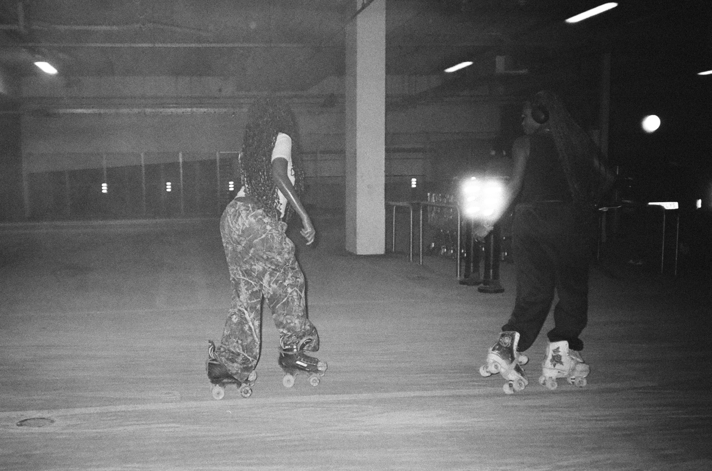 Black and white image of two people roller skating in an indoor rink with concrete pillars and fluorescent lighting overhead.