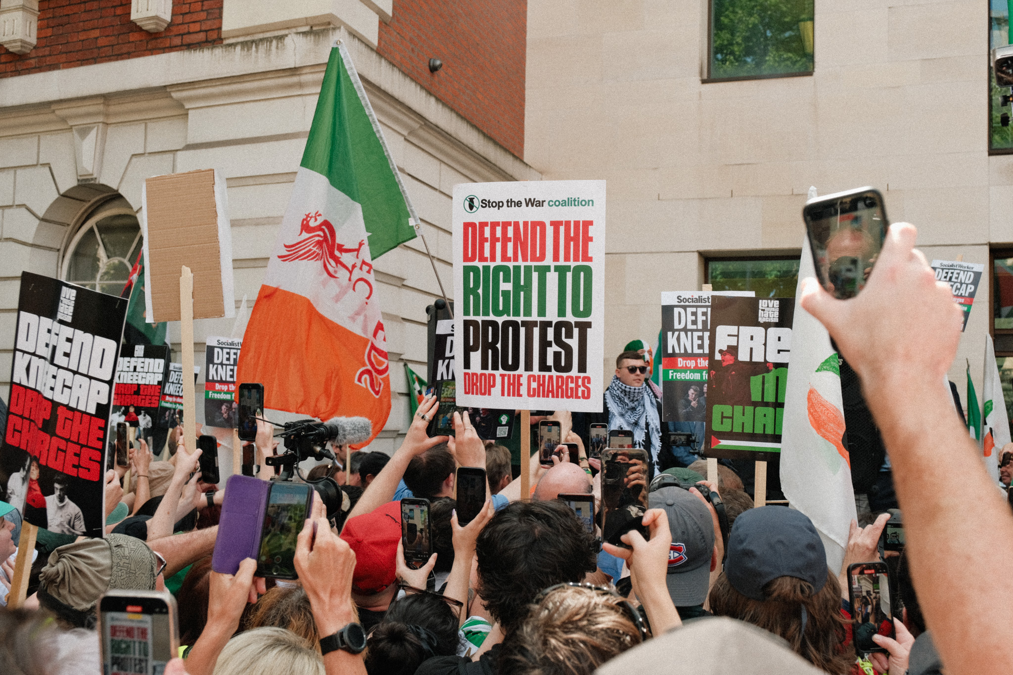 A crowd of protesters holding placards and banners, including flags of Ireland, with "Defend the right to protest" and "Defend knee" written on them.