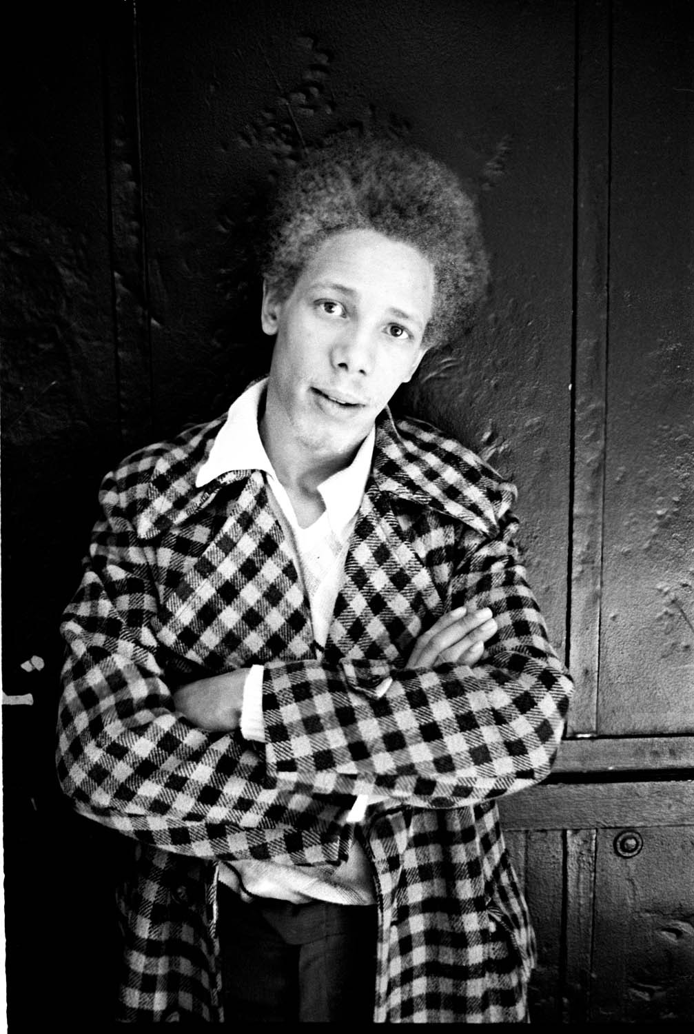 Black and white portrait of person with afro hair wearing chequered coat over white shirt, arms crossed, against wooden door background.