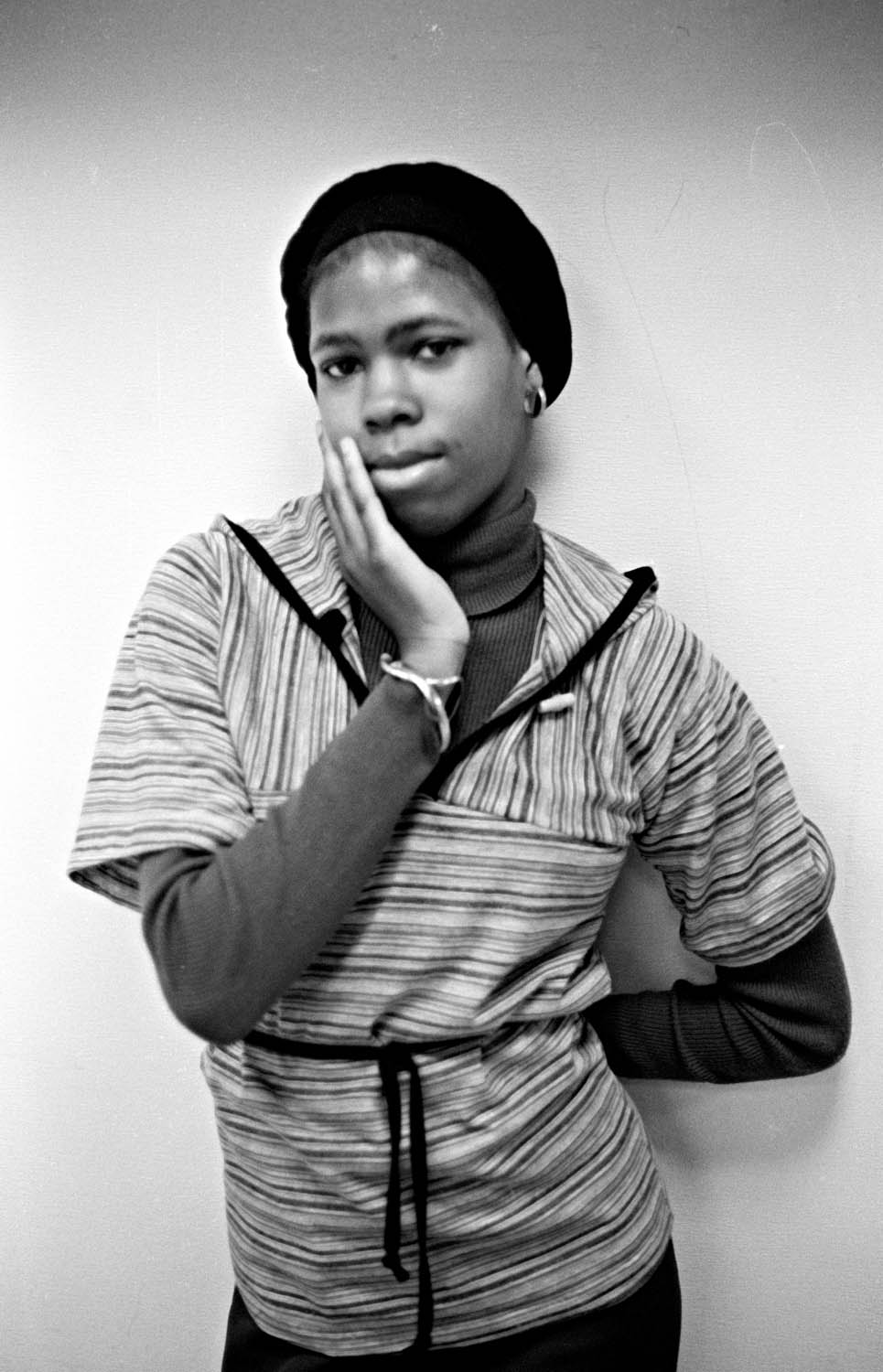 Black and white portrait of person in beret and striped hoodie, hand positioned thoughtfully near face against plain background.