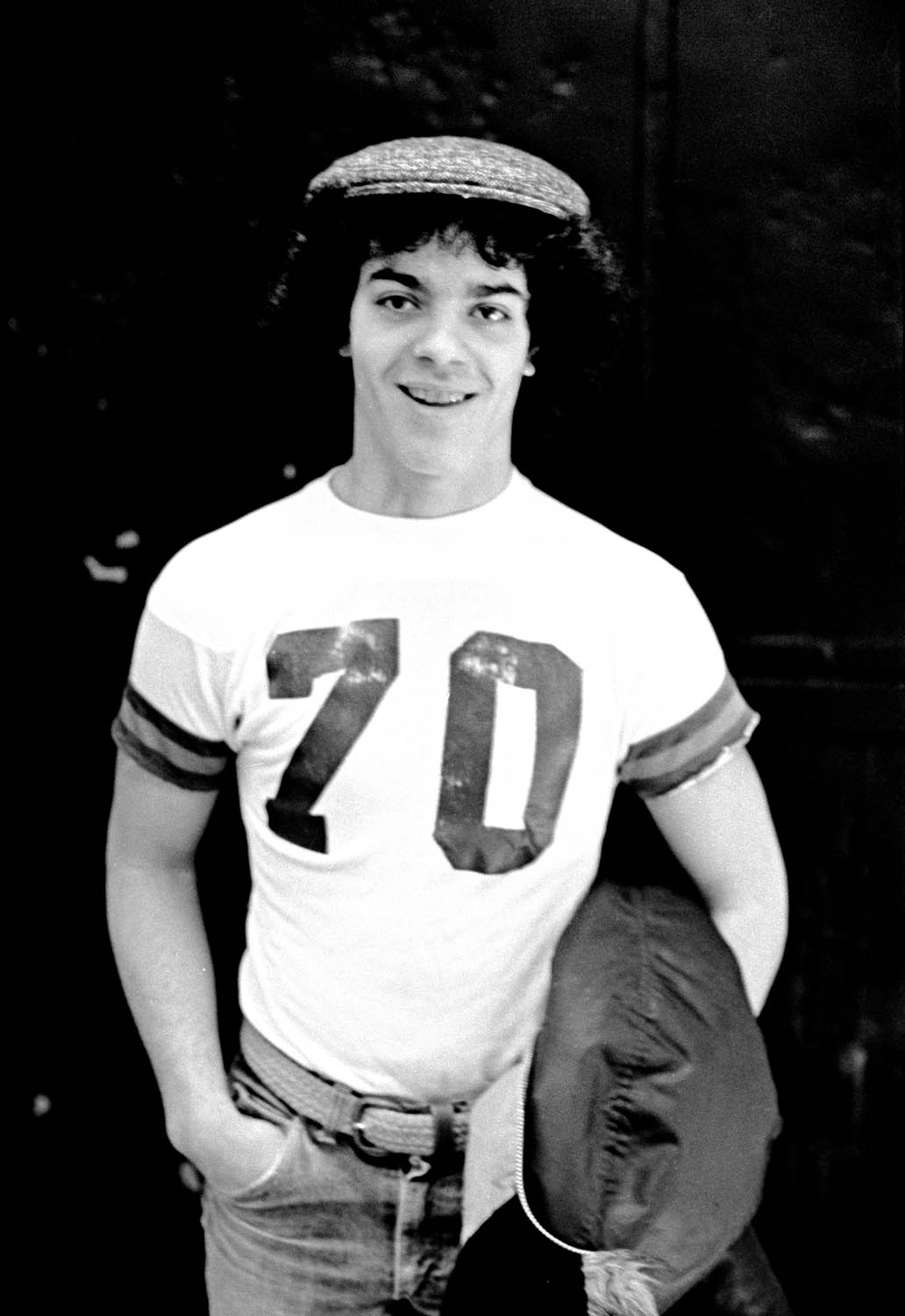 Young man in flat cap and white "70" t-shirt holding American football, smiling at camera in black and white portrait.