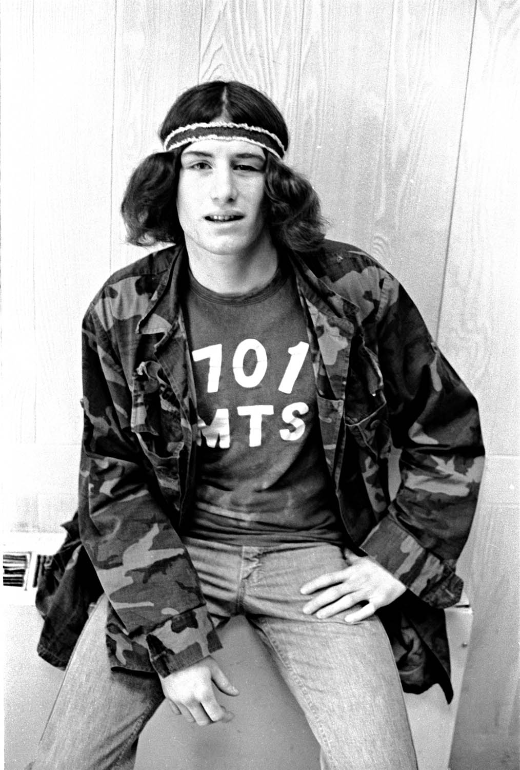 Black and white image of person with shoulder-length hair wearing headband, camouflage jacket over t-shirt, sitting against wooden wall.