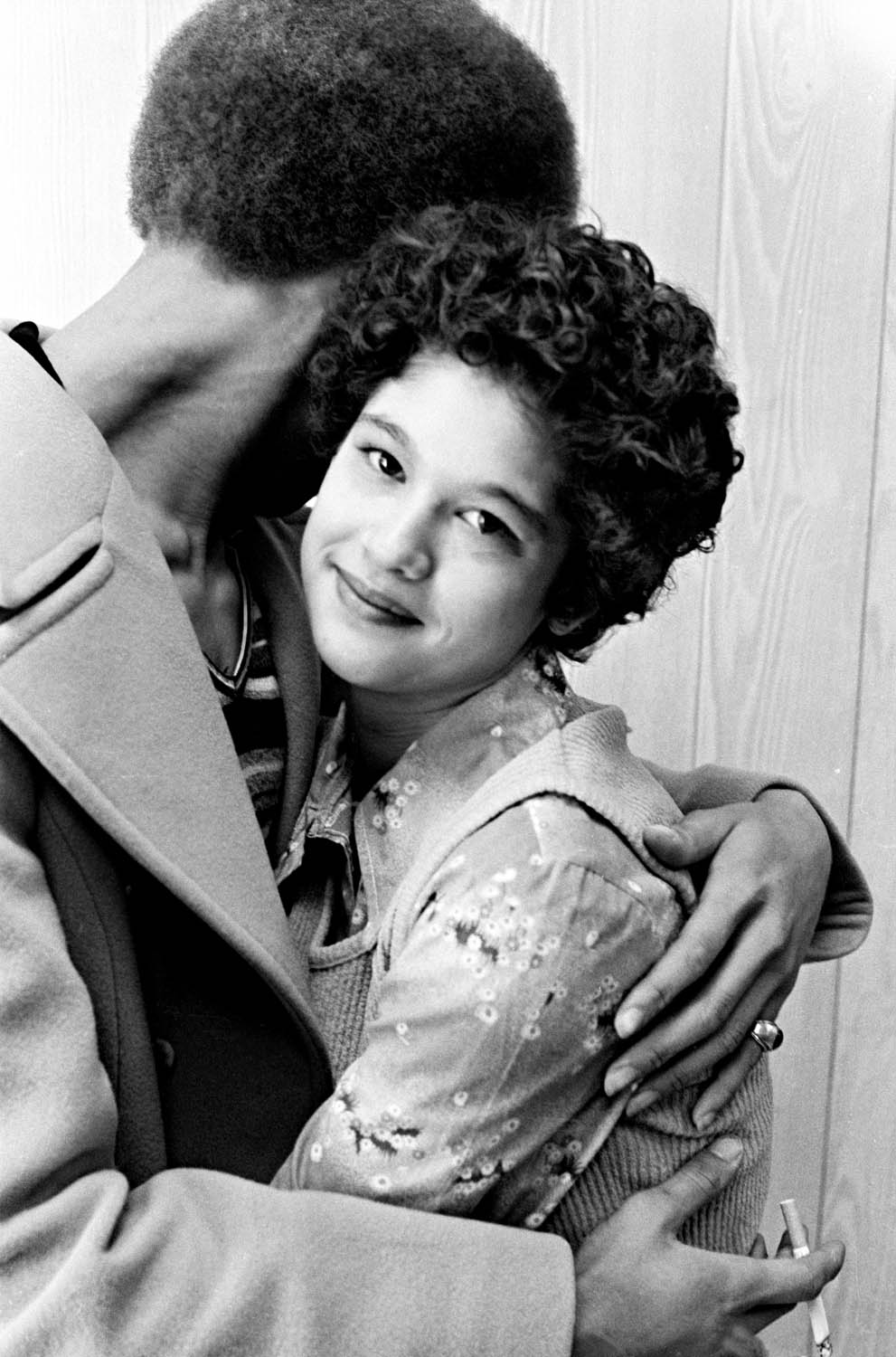 Black and white image of man with afro embracing smiling woman with curly hair wearing floral top, both seated indoors.