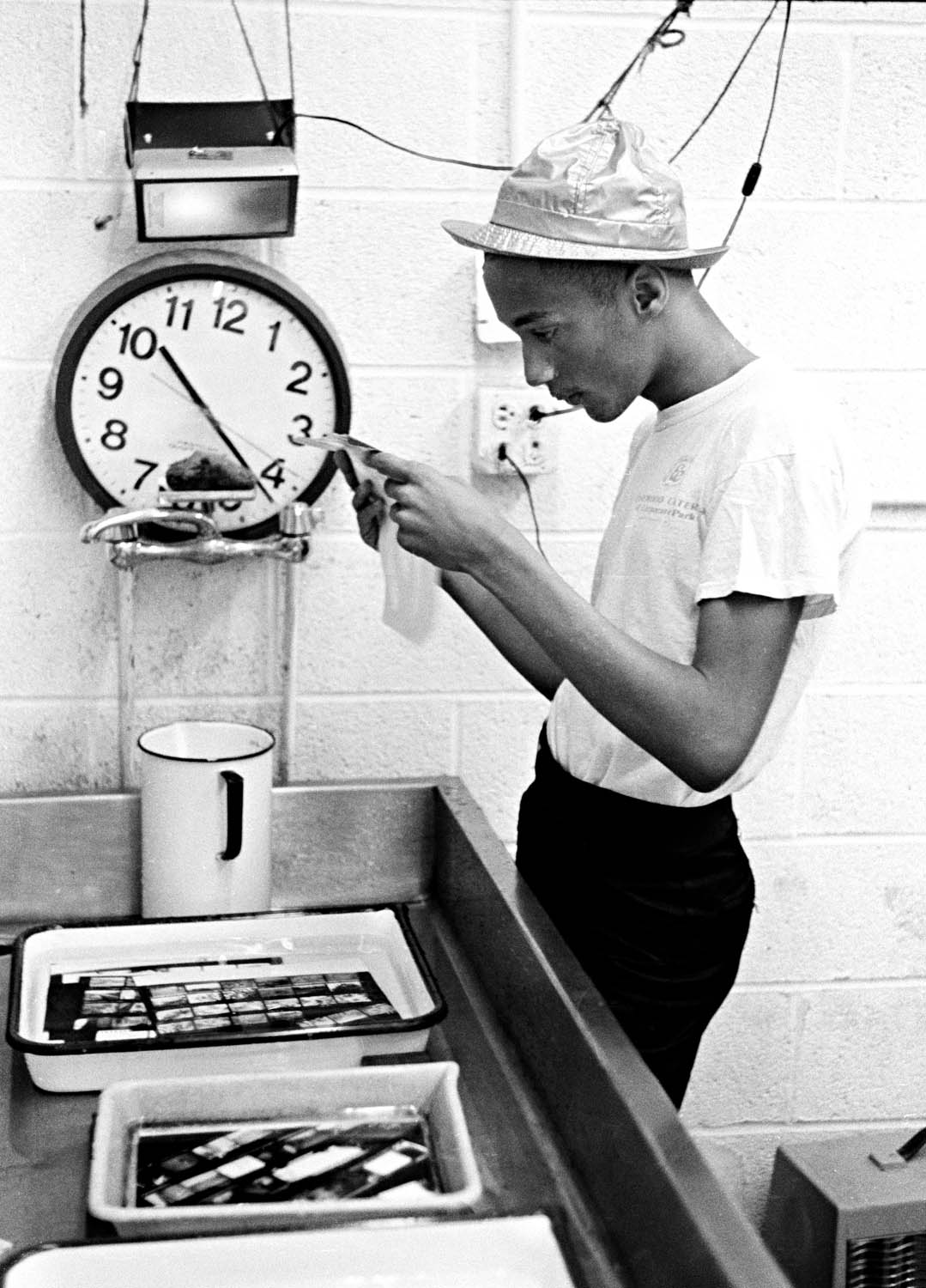 Black and white image: woman in cap using kitchen appliance, wall clock showing 8:40, shelving with containers and trays visible.