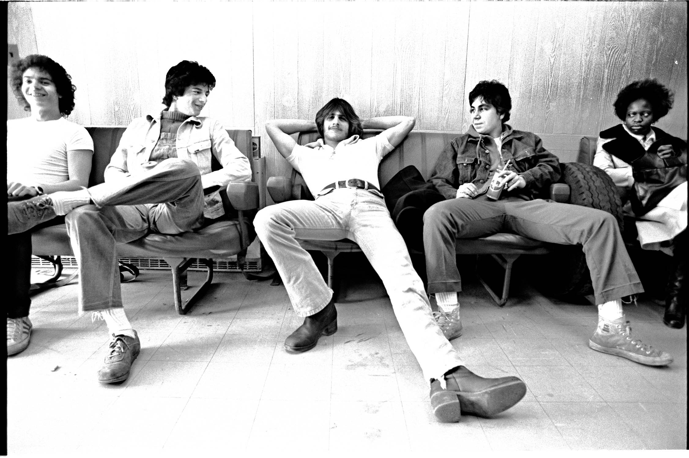 Five people sitting casually on bench seating, black and white image with relaxed poses and casual 1970s clothing.