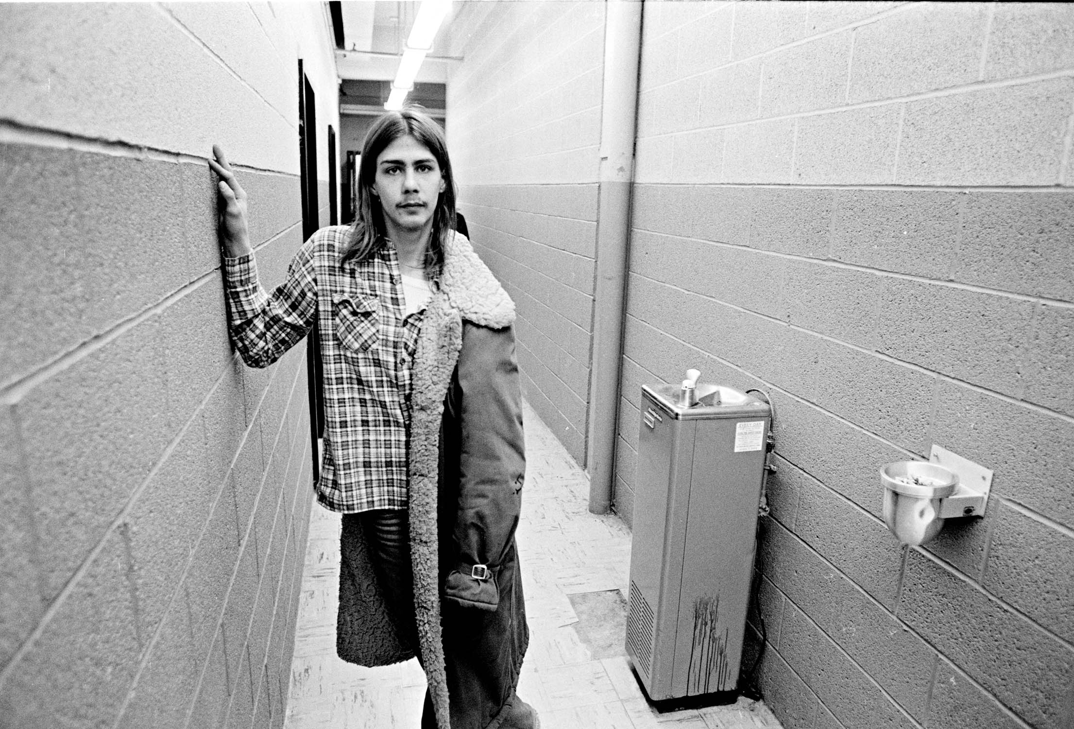 Person in plaid shirt and dark trousers standing in institutional corridor with concrete block walls, fluorescent lighting overhead.