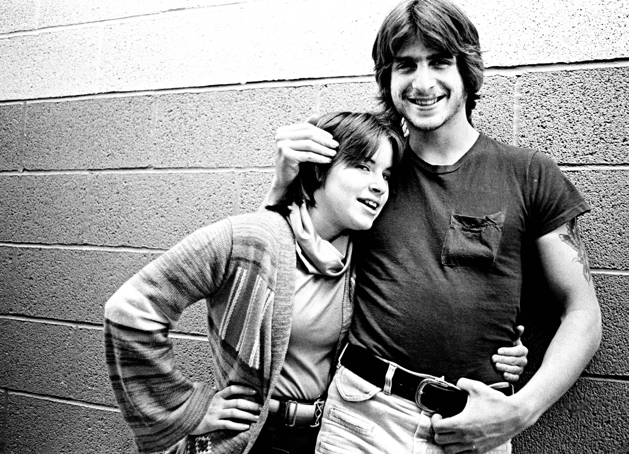 Two young men embracing and smiling against a brick wall, black and white image from what appears to be the 1970s or 1980s.