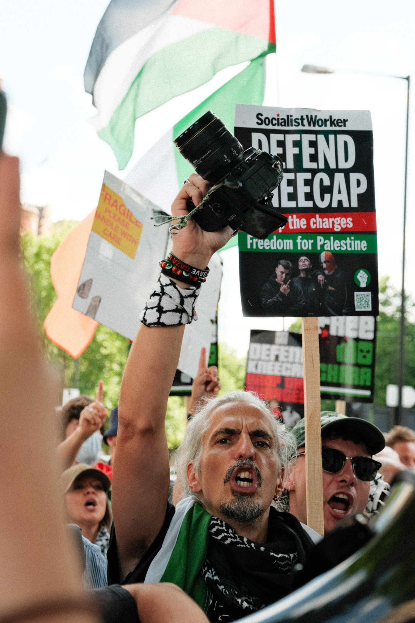 A crowd of protesters demonstrating, with a person holding a "Defend REEACAP" placard and Palestinian flag.