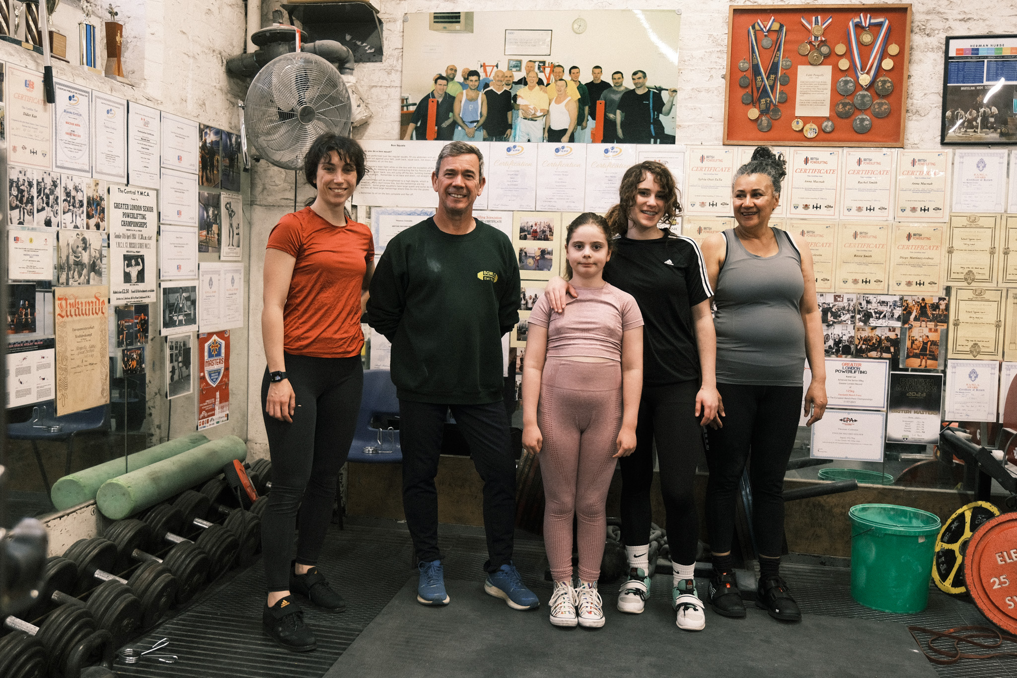 Five people standing in workshop with tools, wall displays, and green equipment. Orange shirt, green shirt, pink outfit, black clothing, grey top.