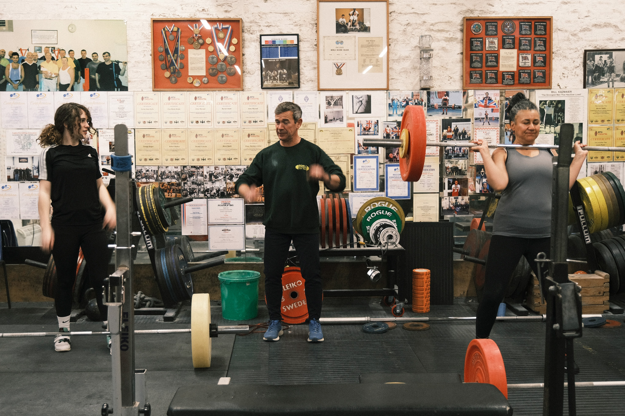 Three people exercising with barbells in gym with white brick walls covered in posters and memorabilia.