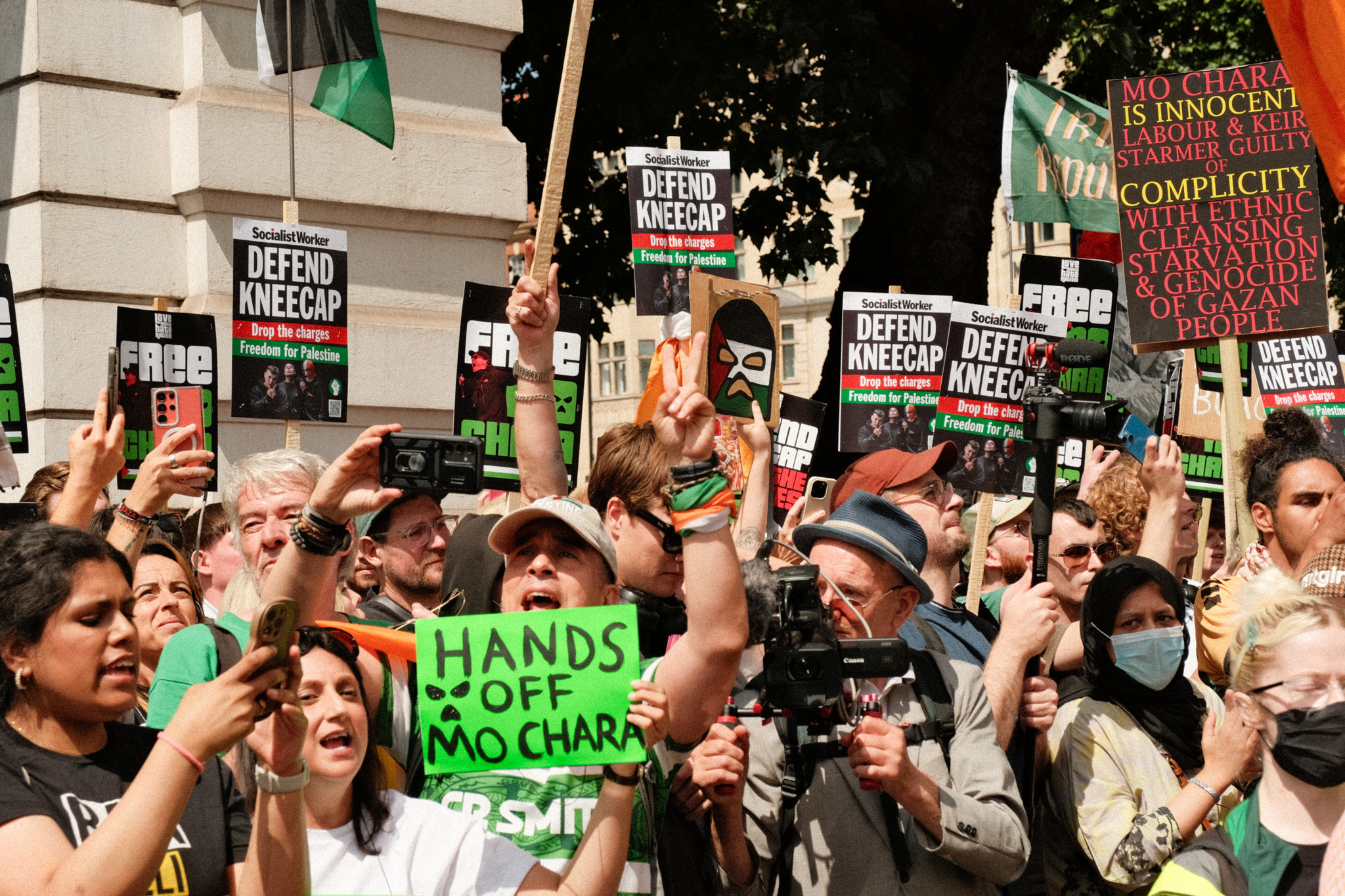 Crowd of protesters holding 'Defend Meecham' placards and 'Hands off Mo Charr' banner, in an urban setting with buildings and trees visible.