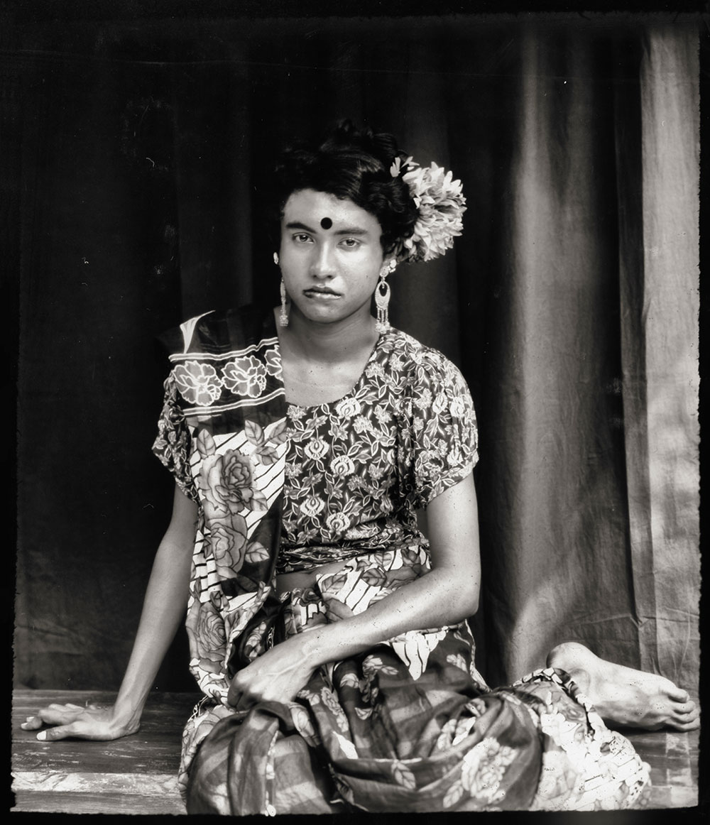 Woman in patterned sari with white flowers in hair, sitting on wooden floor against dark doorway with vertical wooden posts.