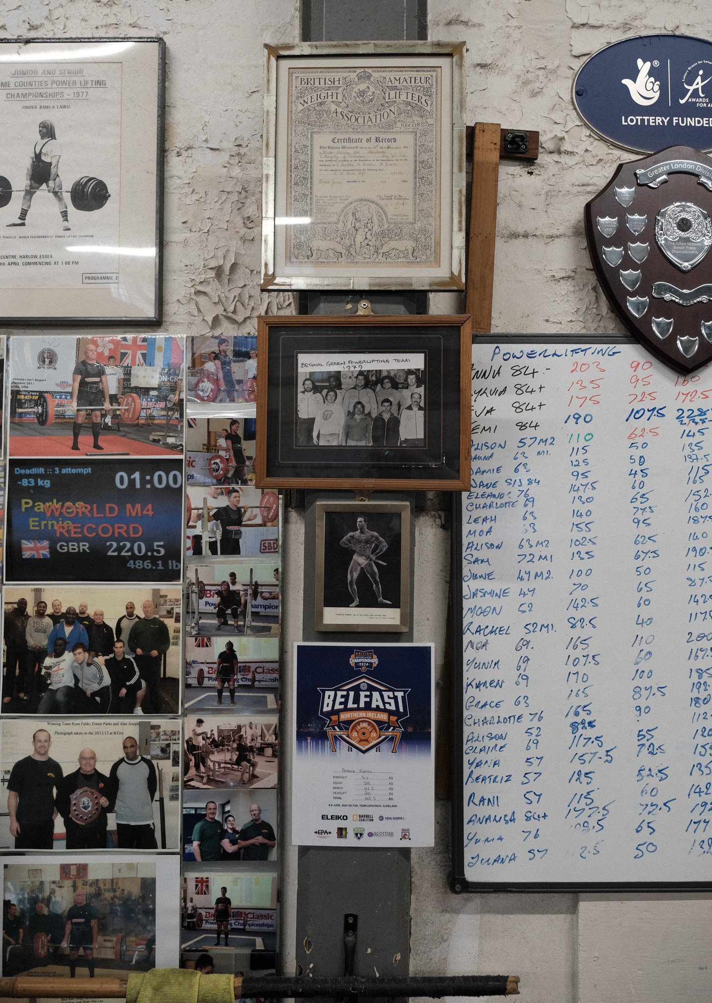 Wall display with framed certificates, boxing photographs, trophy shield, and handwritten scoreboard with blue text on white paper.
