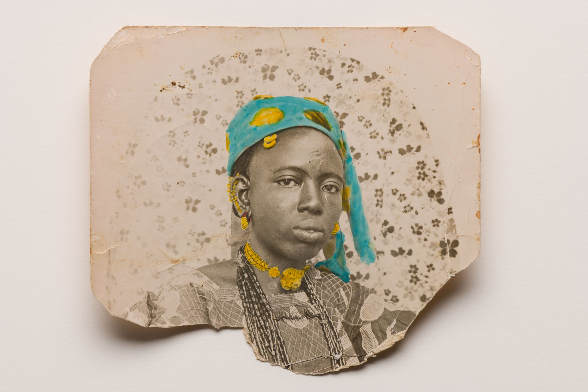 Torn vintage portrait with teal and yellow painted headwrap and jewellery on aged paper with brown spots and stains.