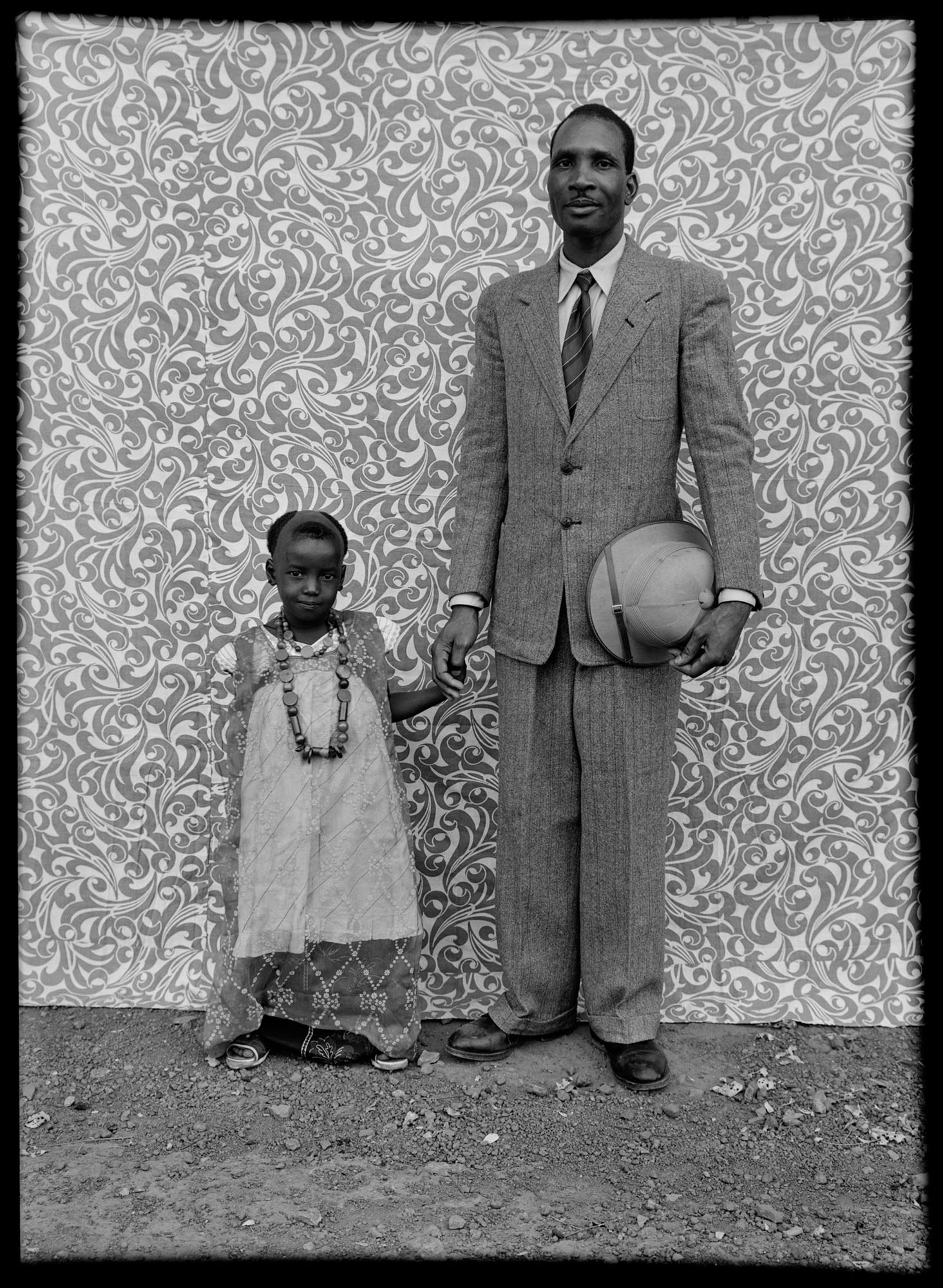 Black and white portrait of tall man in suit holding hat standing next to small child in white dress against ornate patterned backdrop.