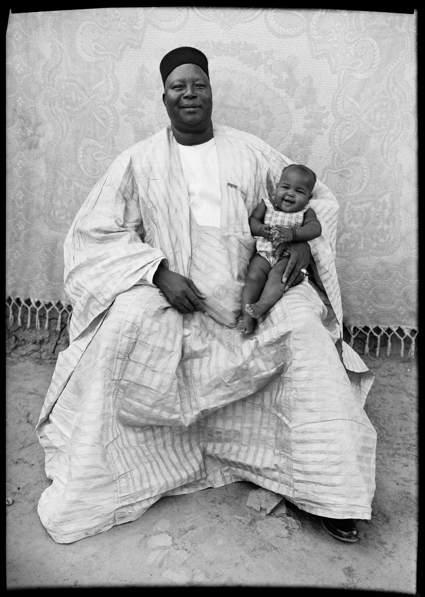 Black and white portrait of man in white traditional robes and cap seated with small child on his lap, both smiling.