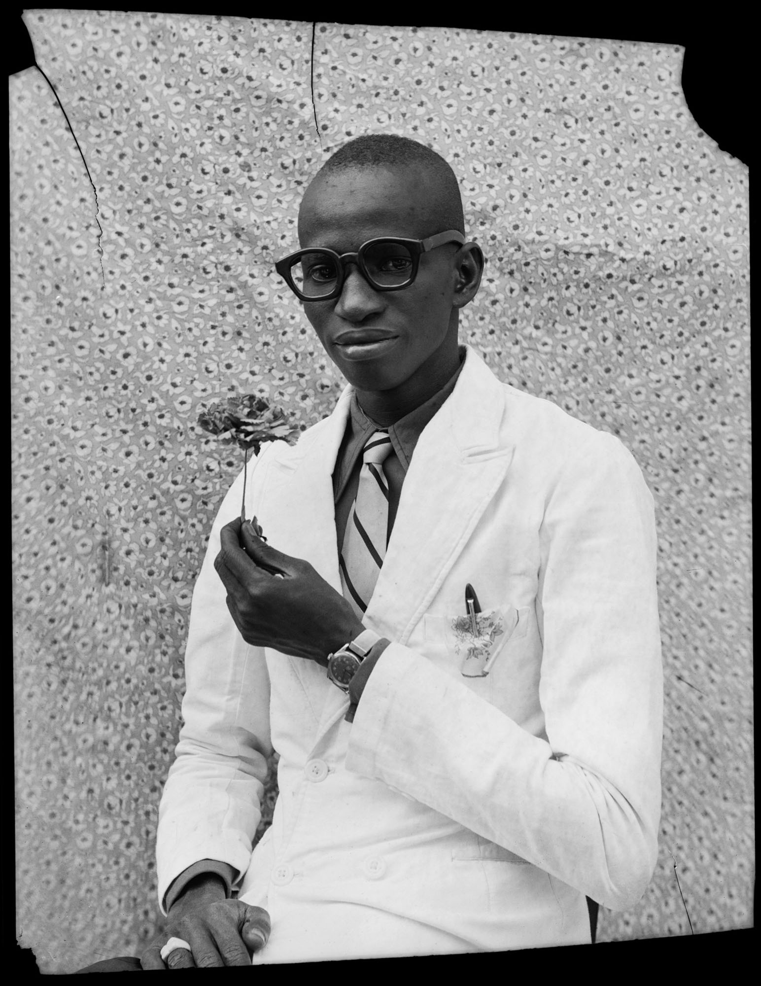 Black and white portrait of man in white coat and glasses holding small plant, against textured wall background.