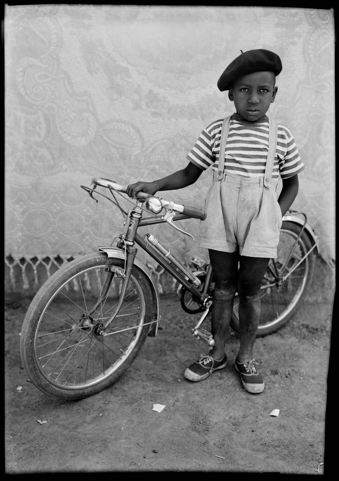 Black and white portrait of young boy in beret, striped shirt, and shorts standing beside bicycle against patterned backdrop.