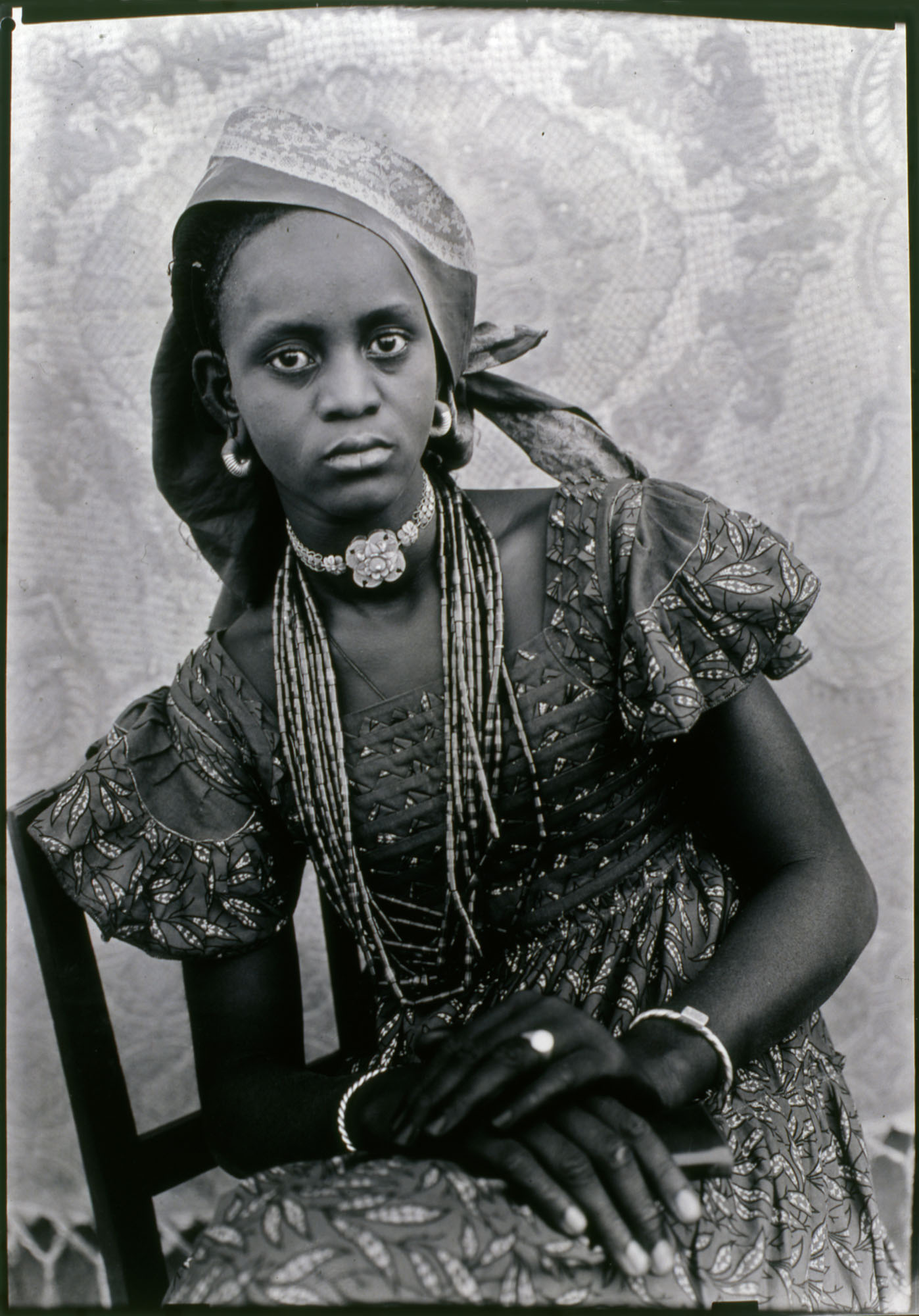 Black and white portrait of woman wearing traditional dress with beaded necklaces, head wrap, and patterned fabric against textured backdrop.