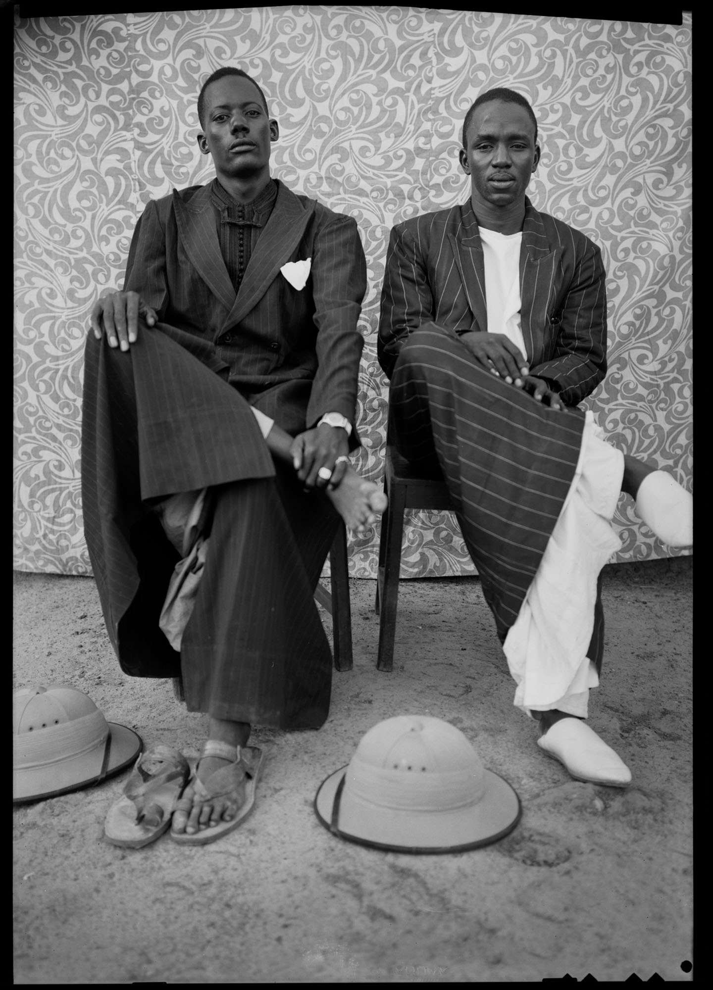 Two men seated on chairs wearing traditional African robes against ornate patterned backdrop. Hat placed on ground. Black and white.