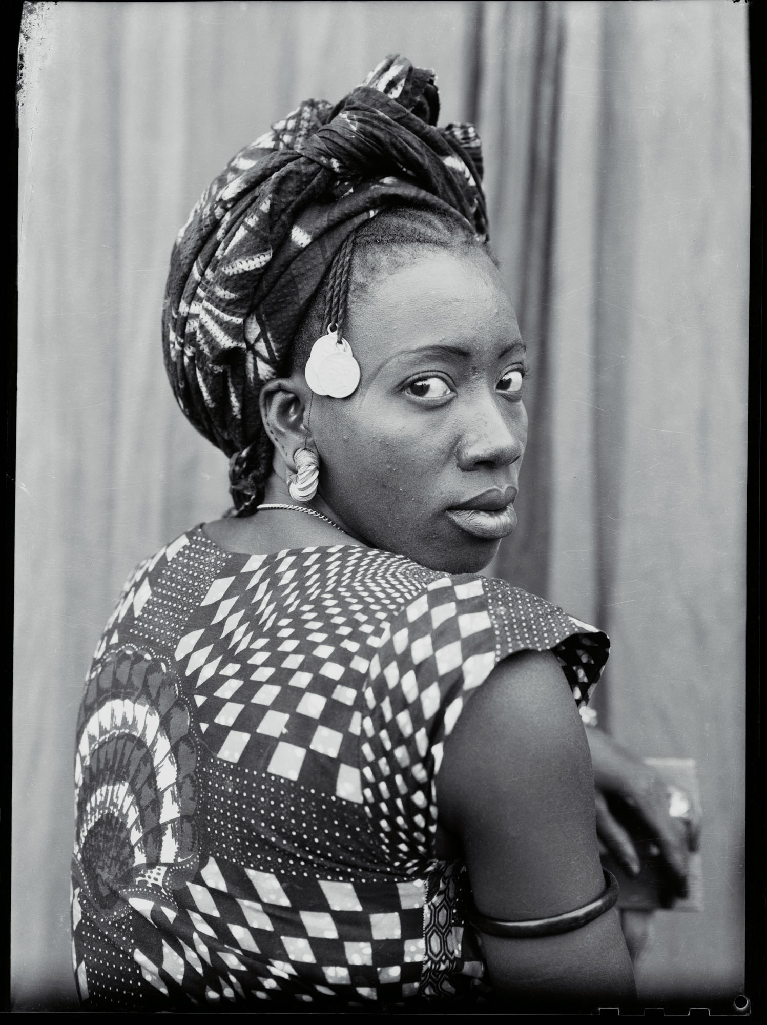 Black and white portrait of woman wearing patterned headwrap and geometric print dress with round earrings, looking over shoulder.