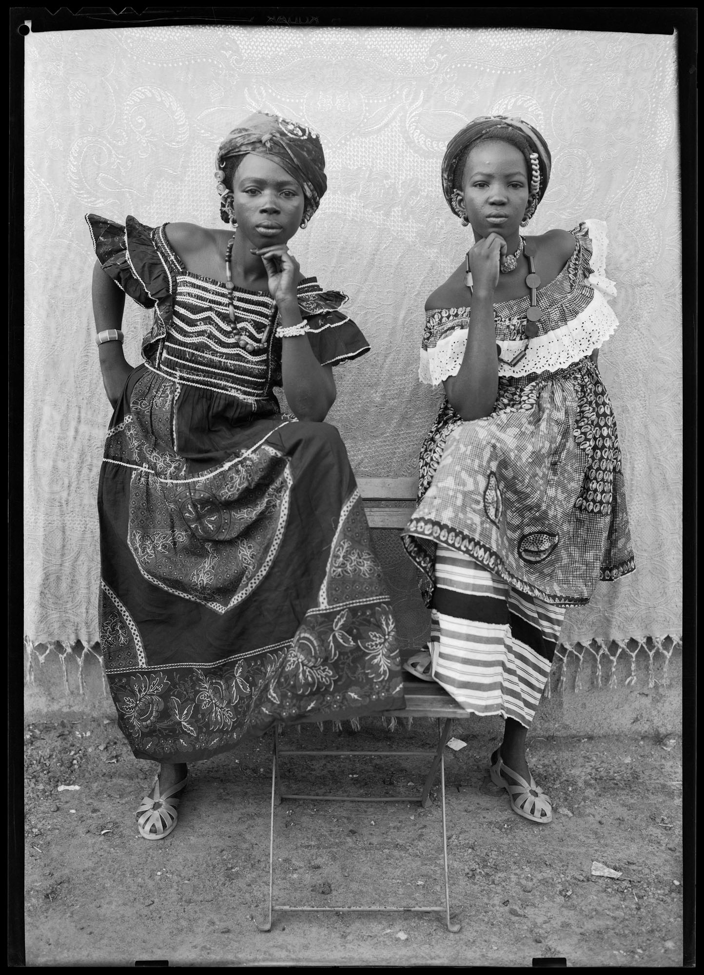 Black and white portrait of two women in traditional African dress with geometric patterns, headwraps, and jewellery, seated on chairs.