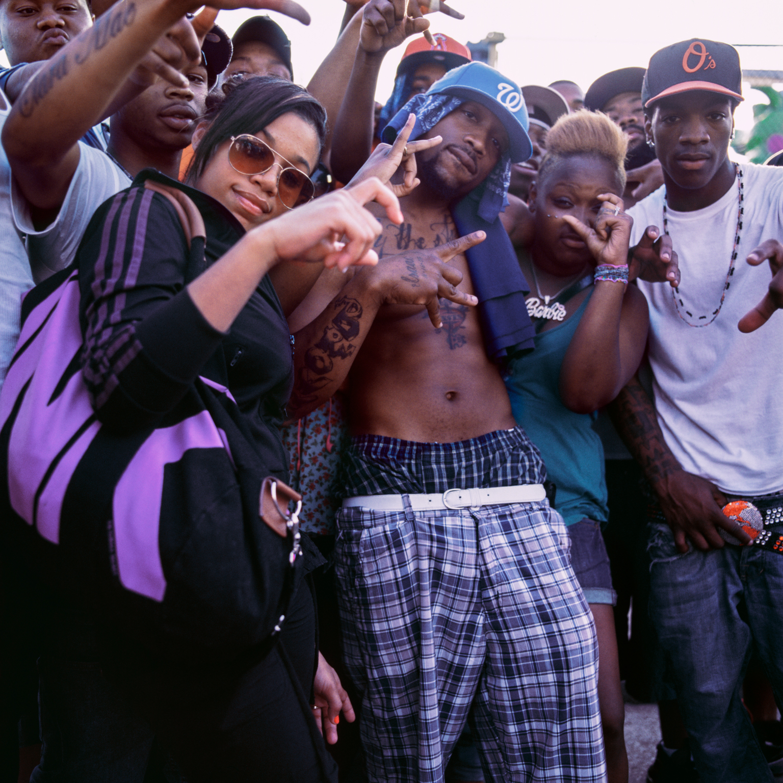 Group of young people making hand gestures at camera, wearing casual clothing including sunglasses, caps, and plaid trousers.