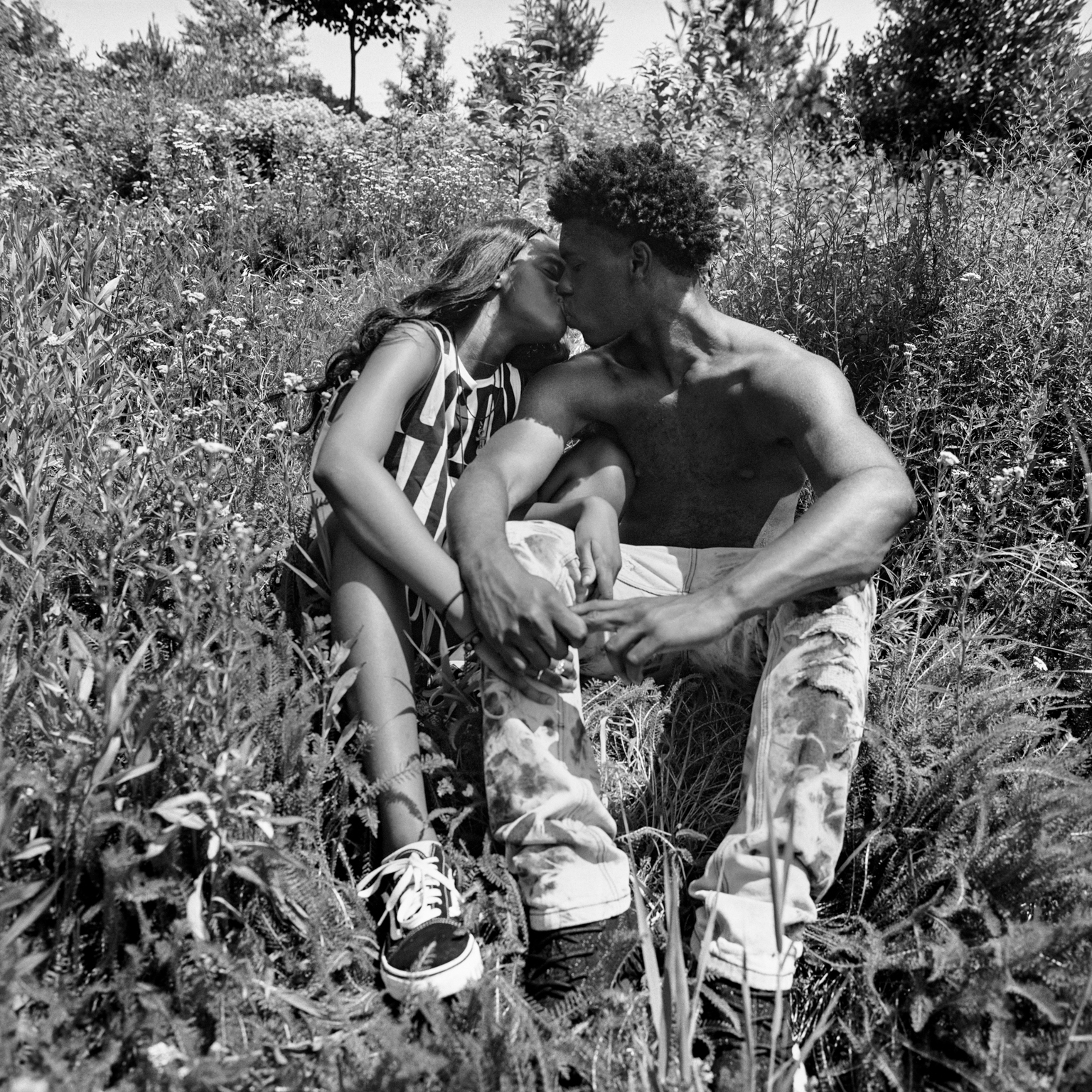 Black and white image of couple embracing whilst sitting in tall grass and wildflowers, surrounded by trees and natural vegetation.