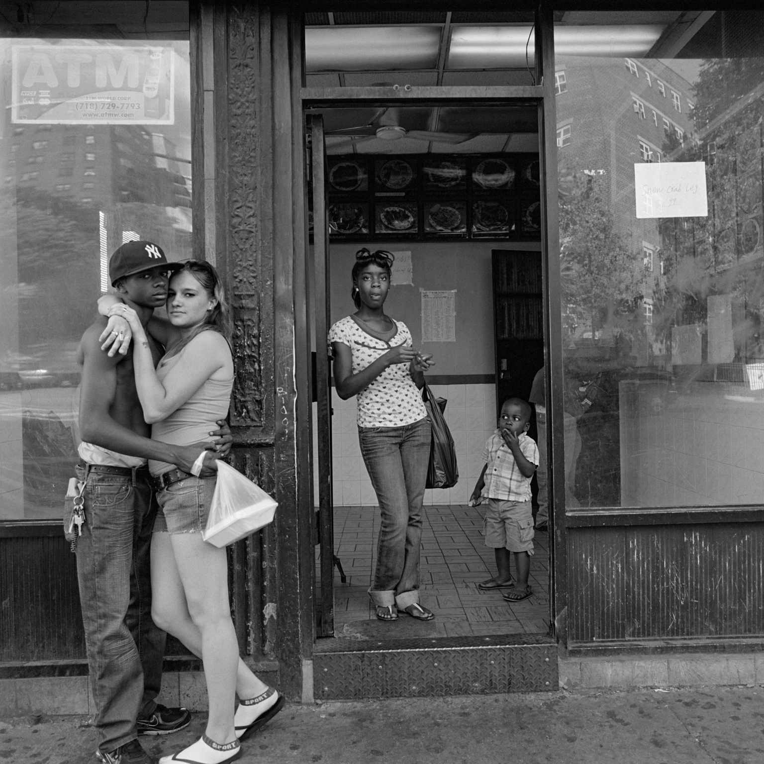 Black and white street scene with couple embracing outside shop, woman in doorway, small child visible, ornate storefront details.