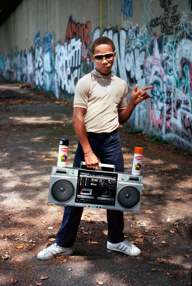 Man in sunglasses and beige shirt holding large silver boombox, standing against colourful graffiti wall with spray paint cans nearby.