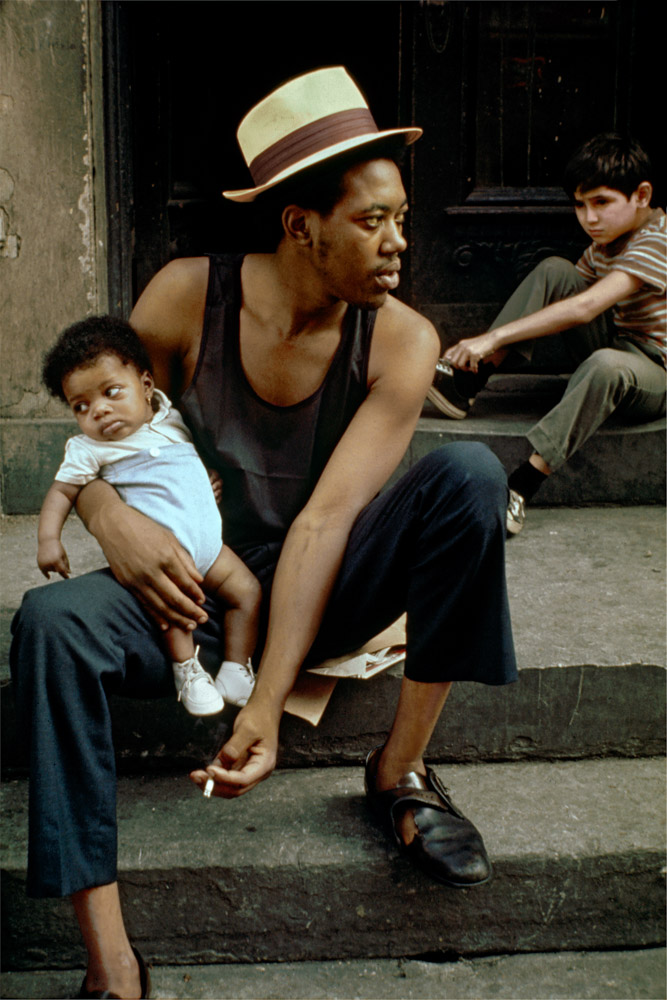 Man in white hat and dark vest sits on steps holding infant, with boy in striped shirt seated nearby on concrete steps.