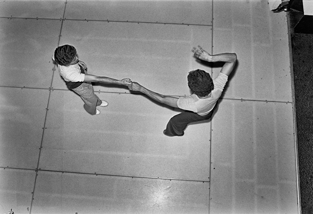 Two swimmers in a pool holding hands whilst floating on their backs, viewed from above in black and white.