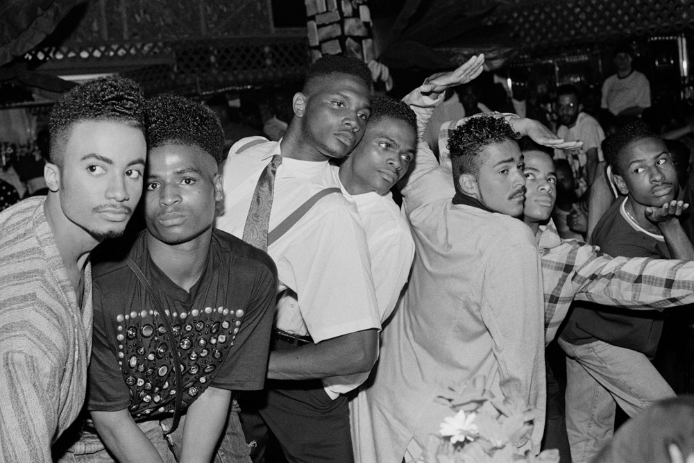 Group of young Black men posing together in crowded indoor venue, black and white image with casual clothing and close positioning.