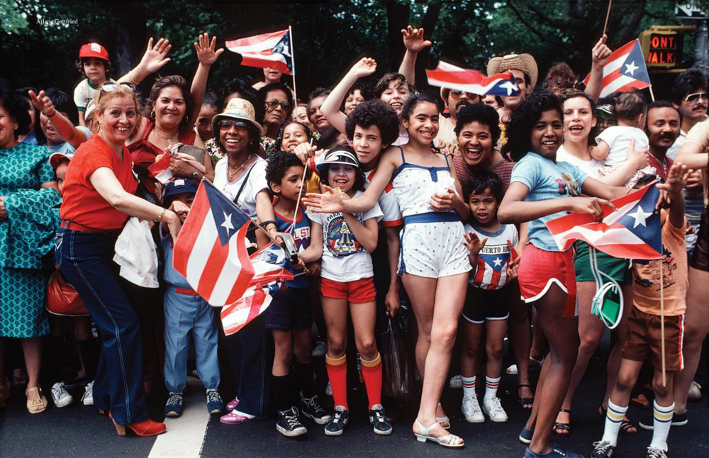 Large group of people holding Puerto Rican flags with red, white, and blue stripes and single star. Crowd celebrating outdoors.