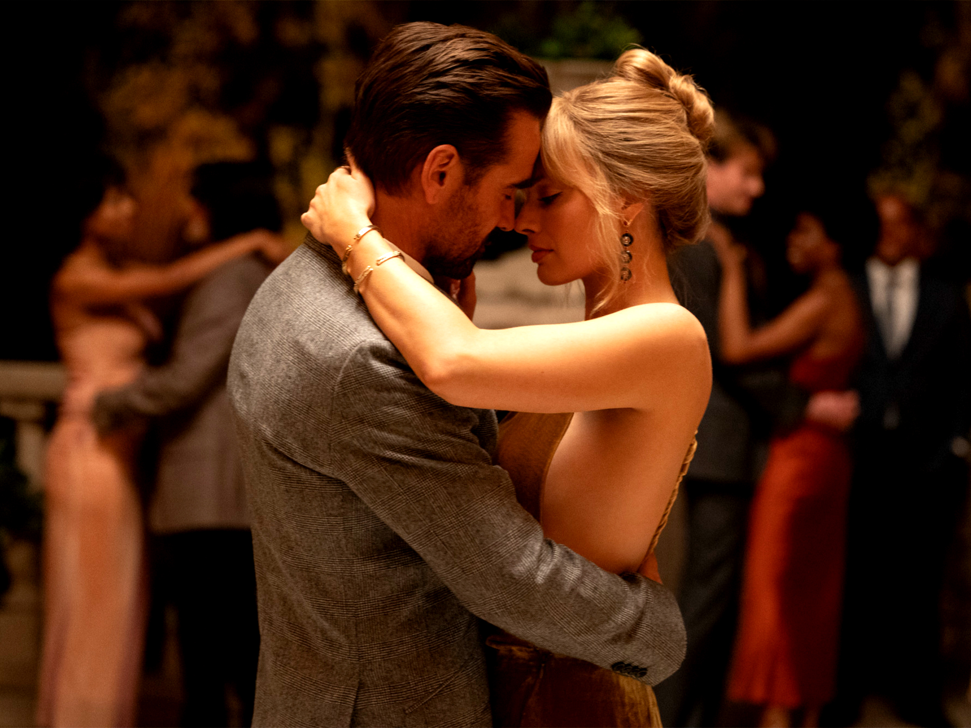 Couple dancing closely, man in grey suit, woman with blonde updo in sleeveless dress, other dancers blurred in background.
